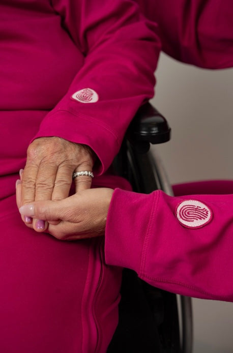 Close-up of two hands gently clasped together, both wearing bright pink sleeves with circular logo patches, seated in a wheelchair.